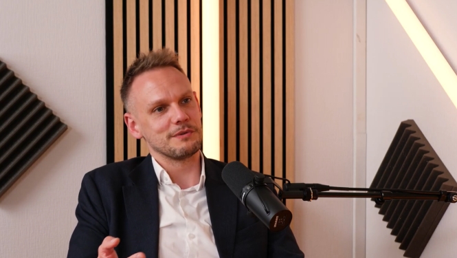 Sekretariāta direktors piedalās podkāsta sarunā, sēžot pie mikrofona skaņu ierakstu studijā. Fonā redzami akustiskie paneļi un koka sienas elementi.