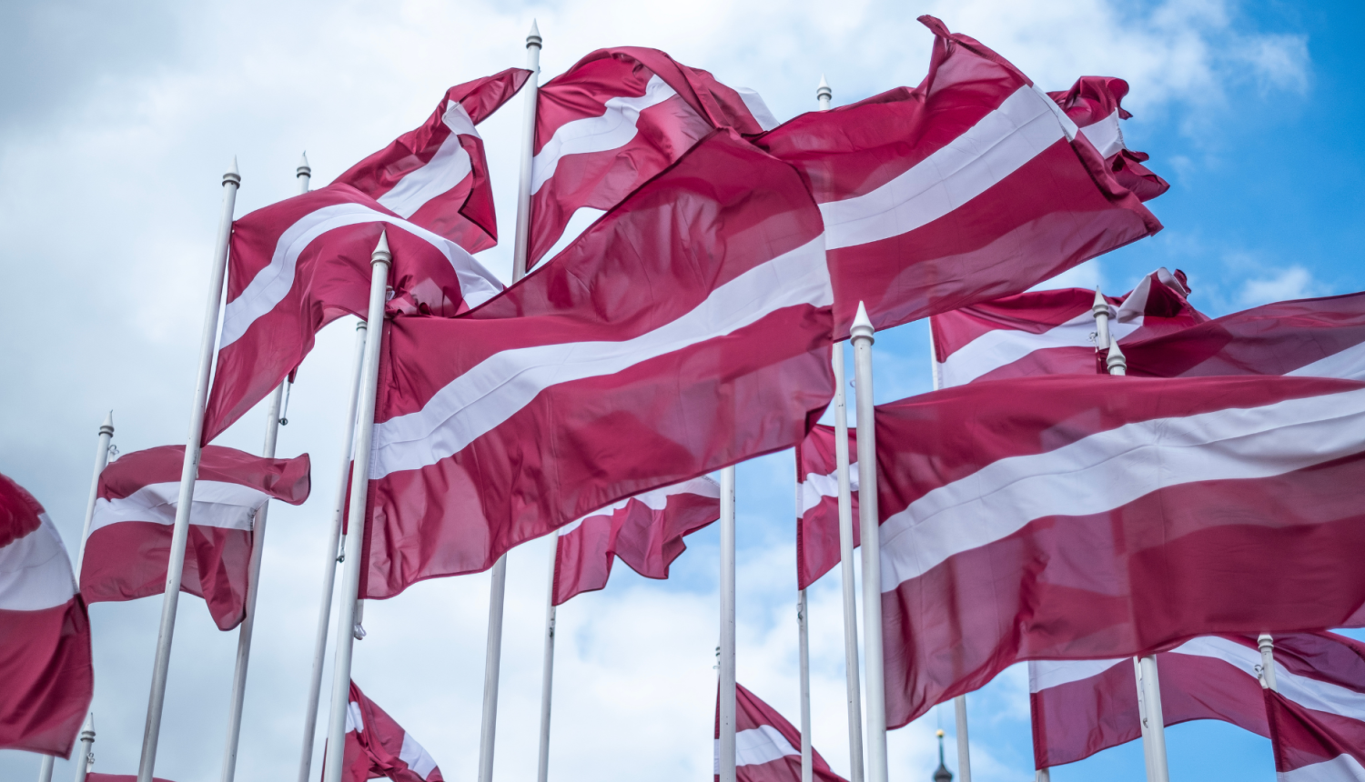 Vējā plīvo vairāki Latvijas valsts karogi uz gaiši zilu debesu fona.