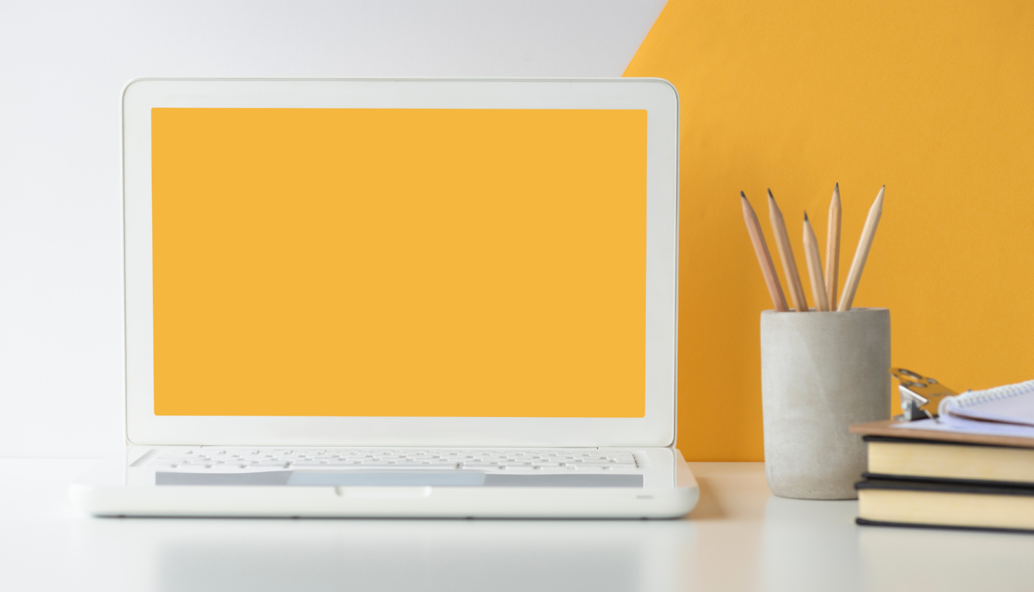 Uz galda ir portatīvais dators ar dzeltenas krāsas ekrānu, blakus tam ir grāmatas un trauks ar zīmuļiem